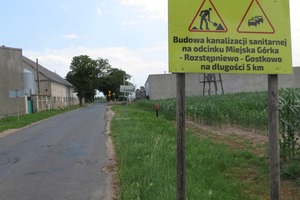 Po prawej stronie zdjęcia widoczna jest żółta tablica z informacją o budowie kanalizacji sanitarnej. Po lewej stronie widoczna jest ulica z zabudowaniami. (photo)