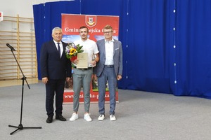 Na zdjęciu znajduje się trzech mężczyzn, pan pośrodku trzyma bukiet kwiatów. (photo)